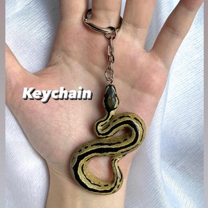 Puede incluir: Un llavero con dise&ntilde;o de serpiente. La serpiente es de color beige con rayas negras y cabeza negra. El llavero incluye un anillo y una cadena de metal. La palabra "Keychain" est&aacute; escrita en texto blanco.
