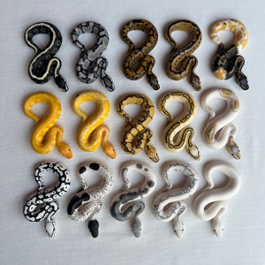 May include: A collection of fifteen handcrafted snake figurines in various colors and patterns. The snakes are arranged in three rows of five, showcasing a variety of designs, including black and yellow stripes, white and gray spots, and solid yellow. Each snake is curled in a stylized pose.