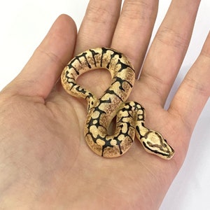 Puede incluir: Una detallada figura de serpiente en miniatura con un patr&oacute;n marr&oacute;n y negro, descansando en una palma abierta. El cuerpo de la serpiente est&aacute; curvado y su cabeza es visible. La figura probablemente est&aacute; hecha de un material duro.