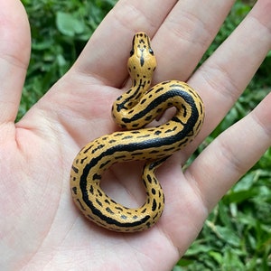 May include: A small, hand-painted wooden snake figurine. The snake has a tan body with black stripes and spots. The head is raised, and the snake is coiled. The figurine is held in an open hand against a green background.