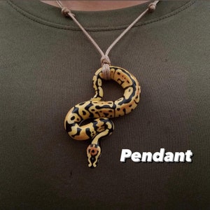 Puede incluir: Un collar con un colgante con forma de serpiente amarilla y negra. El colgante de serpiente cuelga de un cord&oacute;n beige. La palabra "Pendant" est&aacute; escrita en texto estilizado blanco.