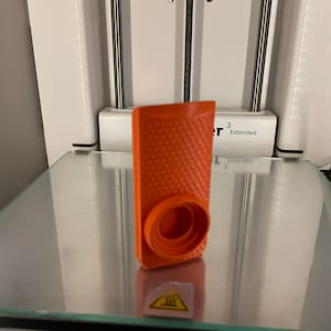 Pode incluir: Um objeto laranja impresso em 3D com uma superfície texturizada e um furo redondo no centro. O objeto está sobre uma superfície de vidro. O texto "Extended" é visível no fundo branco atrás do objeto.
