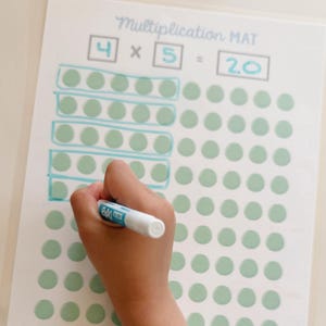 May include: A multiplication mat with the equation 4 x 5 = 20. The mat has a grid of green dots and the equation is highlighted in blue. A hand is holding a white marker, drawing on the mat.