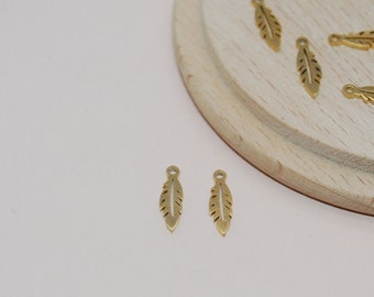 May include: A group of gold-colored metal feather charms on a wooden surface. The charms are small and delicate, with a detailed feather design.