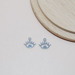 May include: Two small, light blue metal charms in the shape of an eye. Each charm has a detailed eyelash design and a small loop at the top for attaching to jewelry. The charms are on a light-colored wooden surface.