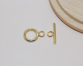 May include: A gold-colored toggle clasp set, featuring a large ring with a small loop, and a straight bar with a loop. The clasp is designed for jewelry making and is shown on a light pink surface.