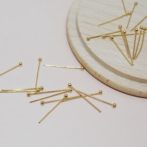 May include: A selection of gold-coloured head pins with small, round ball ends. The pins are scattered on a light pink surface and a wooden cutting board. These pins are likely used for jewellery making or crafting.