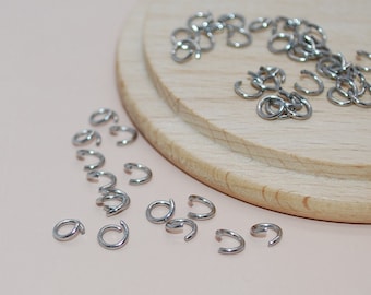 May include: A close-up of a pile of silver jump rings on a wooden surface. The jump rings are small and round, with a gap in the circle. They are used for connecting jewelry pieces.