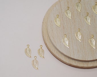 May include: A close-up of a wooden surface with 15 gold leaf charms. The charms are in the shape of a leaf with a small hole at the top. There are three additional leaf charms on a light pink surface.