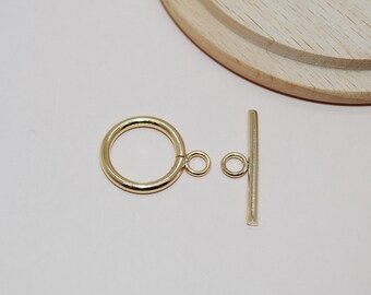 May include: A gold-colored toggle clasp set, featuring a circular ring and a straight bar with small loops for attachment. The clasp is designed for jewelry making and is displayed against a light pink background.