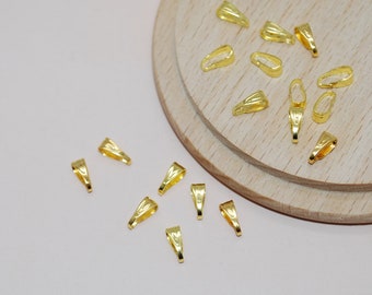 May include: A selection of small, gold-coloured pendant bails scattered on a light wooden surface. The bails are open at the bottom and have a loop at the top for attaching to a chain or cord. The background is a soft, off-white colour.
