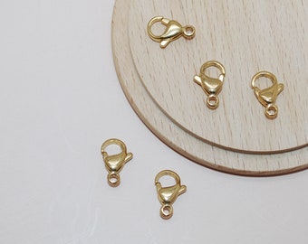 May include: Several gold-colored lobster claw clasps are arranged on a light wood surface. The clasps are small, with a curved hook and a spring-loaded lever. Each clasp has a small loop at the base for attaching to jewelry or other items.