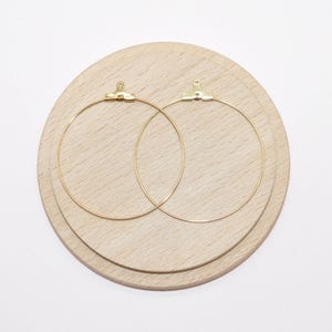 May include: Two gold hoop earrings with a jump ring closure. The earrings are displayed on a wooden surface.