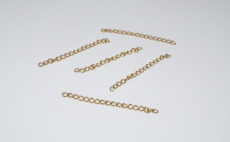 May include: Five gold chain extenders, each with a different length, are laid out on a white surface.