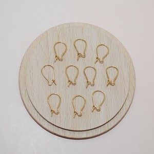 May include: A collection of ten gold-colored earring hooks arranged on a light wooden circular surface. The hooks are in a teardrop shape, with a small loop at the bottom for attaching earrings. The background is a plain white surface.