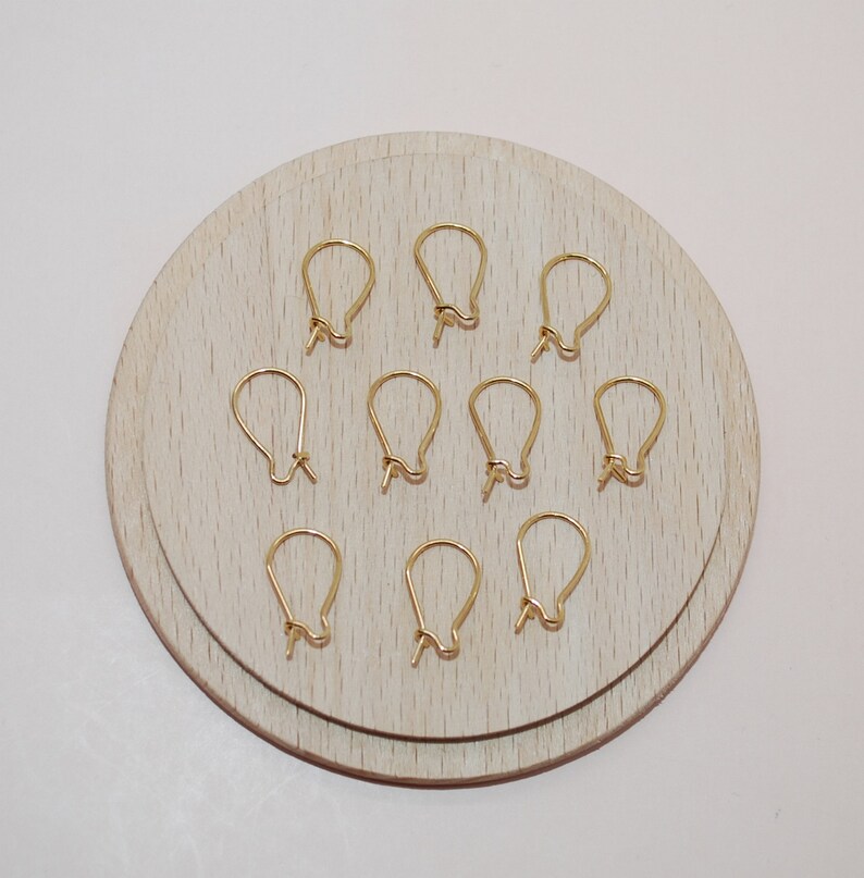 May include: A collection of ten gold-colored earring hooks arranged on a light wooden circular surface. The hooks are in a teardrop shape, with a small loop at the bottom for attaching earrings. The background is a plain white surface.