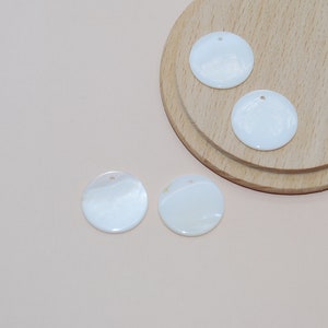 May include: Four white round shell-like sequins with a hole in the center. The sequins are on a light pink surface and a wooden surface.