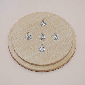 Peut inclure: Cinq breloques en métal argenté de forme circulaire, chacune présentant un motif en étoile en relief. Les breloques sont disposées de manière symétrique sur une surface en bois.