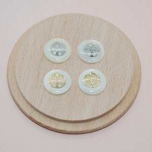 May include: Four white round shell cabochons with gold and silver tree of life designs. The cabochons are displayed on a wooden surface.