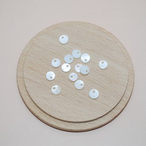 May include: A collection of white, round, mother-of-pearl buttons with a hole in the center. The buttons are arranged on a wooden surface.