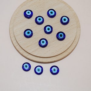 May include: A collection of small, round, blue glass beads with an eye design. The beads are arranged on a light wooden surface and a pale pink background. The eye design features a black pupil, a light blue iris, and a dark blue outer ring.