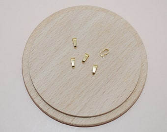 May include: A selection of small, gold-coloured jewellery findings, including several rectangular bails and a single oval clasp. The findings are arranged on a light-coloured, round wooden surface, set against a soft pink background.