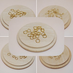 以下が含まれることがあります： 木製の表面に置かれた金色のジャンプリングの山をクローズアップした写真。ジャンプリングは小さく丸く、ジュエリー作りに最適です。