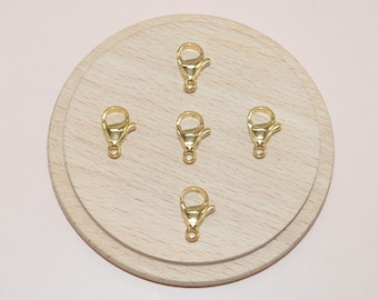May include: Five gold-colored lobster claw clasps arranged on a light wooden surface. The clasps are evenly spaced and have a shiny finish. The background is a soft pink.