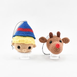 Op de afbeelding: Twee gehaakte kerstversieringen: een elf met een blauwe, rode en gele hoed, en een rendier met bruine geweien en een rode neus. Beide hebben zwarte ophangkoorden en staan op transparante standaards.
