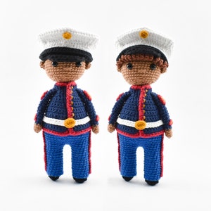 Könnte beinhalten: Zwei gehäkelte Puppen, die als Marines verkleidet sind. Beide Puppen haben braune Haut, schwarze Haare und tragen blaue Uniformen mit roten Verzierungen und einem weißen Gürtel. Eine Puppe hat einen schwarzen Hut und die andere einen braunen Hut.