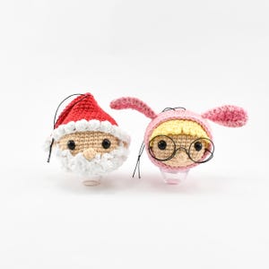 May include: Two crocheted ornaments, one is a Santa Claus with a white beard and the other is a person with blonde hair wearing pink bunny ears and glasses.