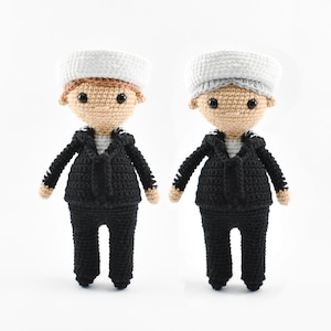 May include: Two crocheted dolls dressed in black sailor outfits with white hats. The dolls have brown yarn hair and black eyes.