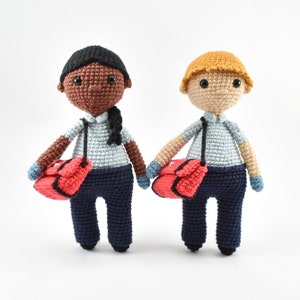 Könnte beinhalten: Zwei gehäkelte Puppen, die blaue Hemden und marineblaue Hosen tragen. Beide Puppen tragen rote Taschen mit weißen Akzenten. Eine Puppe hat braune Haut und schwarze Haare, die andere Puppe hat helle Haut und blonde Haare.