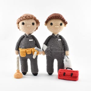 Może przedstawiać: Dwie szydełkowe lalki ubrane w stroje budowlańców. Jedna lalka trzyma tłok i pas narzędziowy, druga trzyma puszkę farby i skrzynkę z narzędziami.