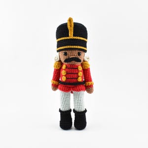 May include: A handmade crochet nutcracker doll. The doll is dressed in a red jacket with gold buttons, black trousers, and a black hat with gold accents. The doll has a brown face, a black moustache, and white hair.