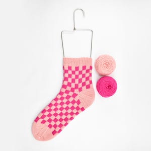 May include: A pink and white checkered sock hanging on a metal hanger with two balls of pink yarn.