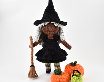 May include: A crocheted witch doll with black hair and a black dress, holding a brown broom. The doll is standing next to three crocheted pumpkins, one of which is a trick-or-treat bucket.