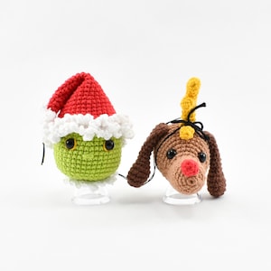 Puede incluir: Dos adornos navideños de crochet, uno verde con un gorro de Papá Noel rojo y el otro marrón con una cuerna de reno amarilla.