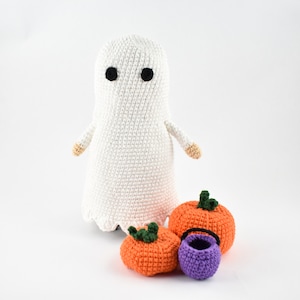 May include: A white crocheted ghost with black eyes stands next to three crocheted pumpkins. Two of the pumpkins are orange and one is purple. The purple pumpkin is a trick-or-treat bucket.