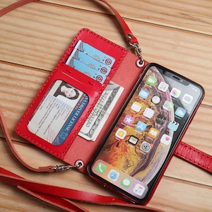 Puede incluir: Una funda de teléfono de cuero rojo con una billetera adjunta. La funda tiene una ranura para una licencia de conducir y tarjetas de crédito. La funda del teléfono tiene una correa para llevarla alrededor del cuello.