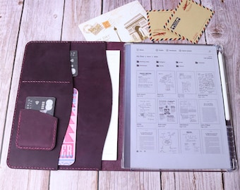 May include: A burgundy leather portfolio with a clear plastic pocket for a tablet or notebook. The portfolio has multiple pockets and slots for cards, pens, and other accessories. The portfolio is open and a white stylus is visible in the clear pocket.