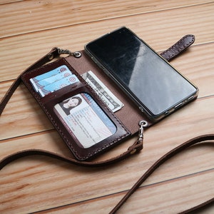 May include: A brown leather phone case with a strap. The case has slots for cards and cash. A smartphone is inside the case.