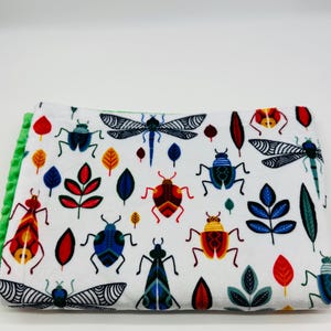 May include: White baby blanket with a colorful insect and leaf pattern. The blanket features various stylized bugs in red, blue, yellow, and green, along with matching leaves. The edge is trimmed with a green plush material.