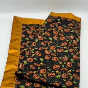 Può includere: Una copertina per neonati nera e arancione con un motivo a zucca e un bordo in raso dorato.