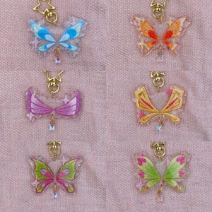 Könnte beinhalten: Sechs bunte schmetterlingsförmige Anhänger mit goldfarbener Hardware und hängenden Kristallakzenten. Die Flügel sind in Blau-, Orange-, Rosa-, Gelb- und Grüntönen gehalten. Jeder Anhänger hat einen sternförmigen Akzent.