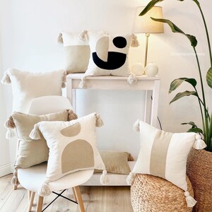 May include: Five beige throw pillows with tassels, two with geometric patterns, one with a black and white geometric pattern, and two with a single beige stripe. The pillows are arranged on a white chair, a white table, and a woven stool. A lamp with a beige shade is on the table. A potted plant is in the background.
