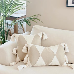 May include: Two beige and white diamond patterned throw pillows with white tassels on a white couch.