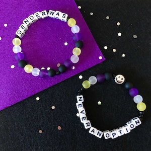 Könnte beinhalten: Zwei Perlenarmbänder mit schwarzen, gelben und violetten Perlen. Das erste Armband hat weiße Buchstabenperlen, die "GENDER WAS NEVER AN OPTION" buchstabieren. Das zweite Armband hat eine Smiley-Perle und weiße Buchstabenperlen, die "NEVER AN OPTION" buchstabieren.
