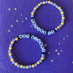 Puede incluir: Dos pulseras de cuentas amarillas y moradas. Una pulsera deletrea "NATURE IS" y la otra "NOT BINARY".