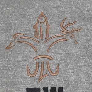 Pode incluir: Uma bolsa de tecido cinza com um desenho bordado marrom de um lírio com cabeças de peixe, pato e veado. A bolsa tem as letras "TW" bordadas em preto abaixo do desenho.
